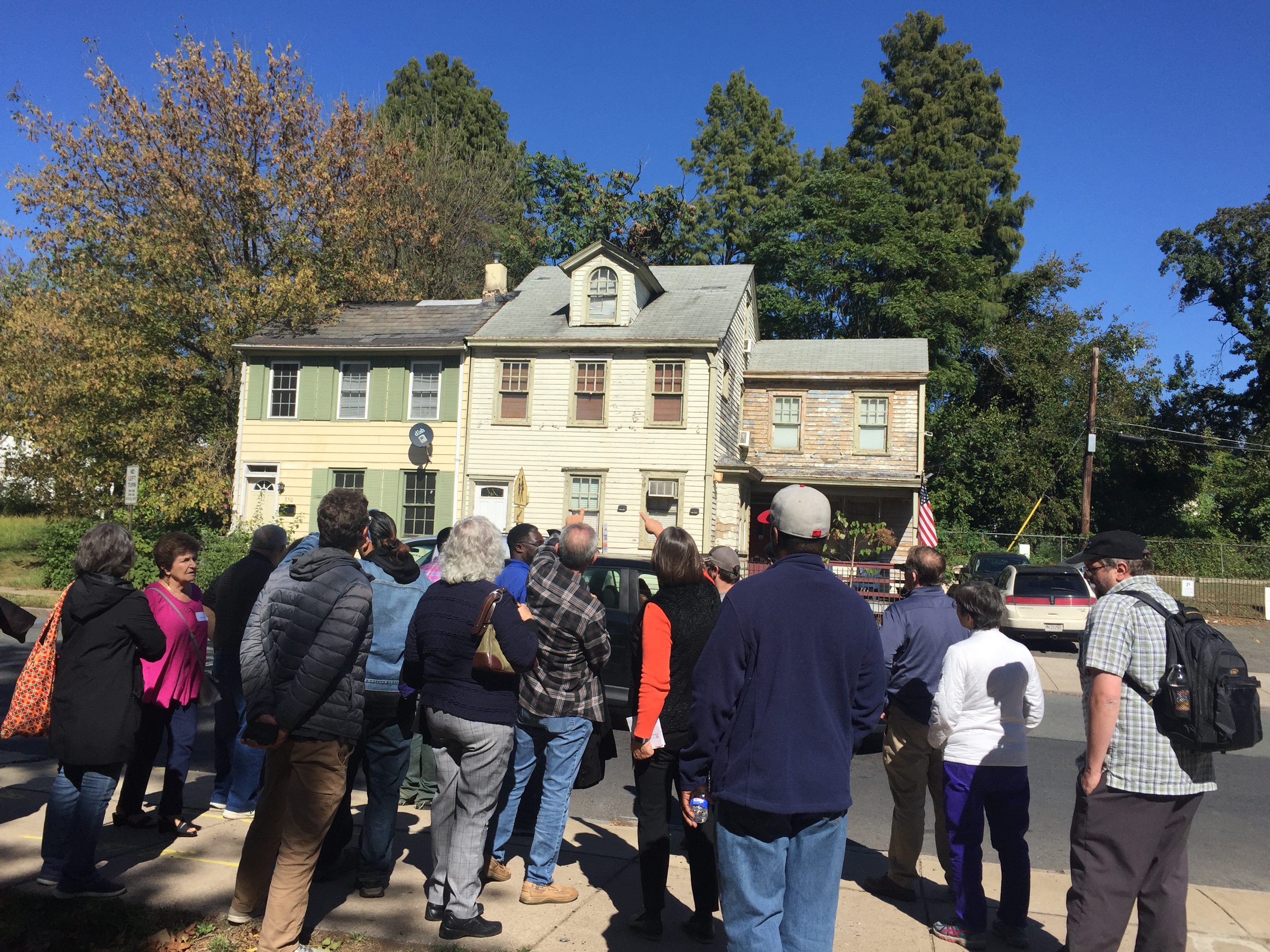 This is a color photograph of a group of people taking walking tour looking at historic buildings in Trenton, NJ. They are pointing toward one of two buildings sided in wood in the background.