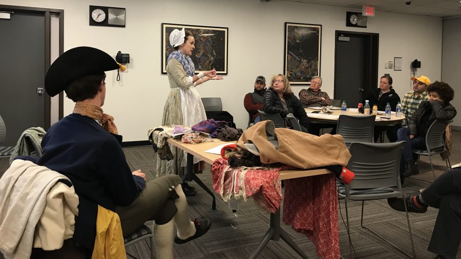 In this color photograph, a man and a woman dressed in repdocution eighteenth-century costume talk to participants sitting at tables.